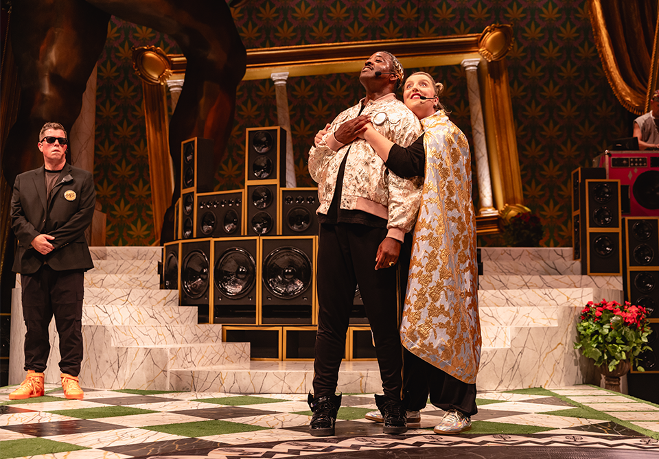 Three people on a theatrical stage with marble steps and stacked speakers. Two stand closely together in the center, with one draped in a cape, and a suited person stands on the left.