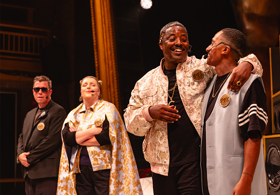 Four people on stage, two on the left in dark attire, and two on the right smiling with light-colored jackets.