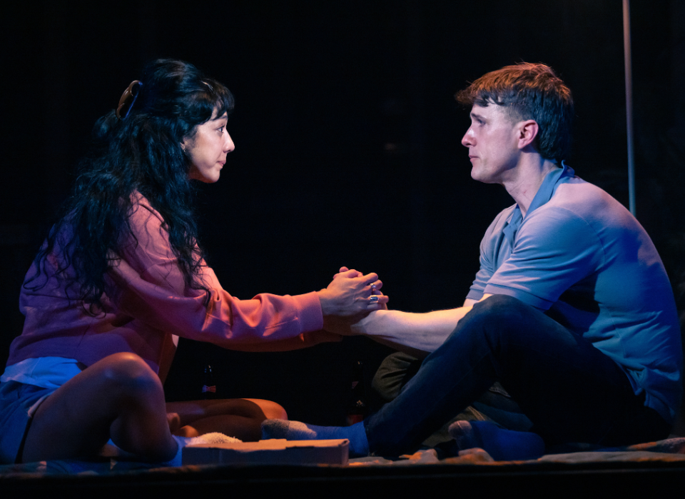 Two people sit facing each other on the floor, holding hands, under dim lighting.