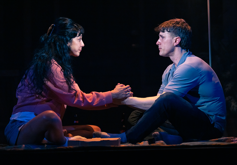 Two people seated, facing each other, holding hands in a dimly lit setting.