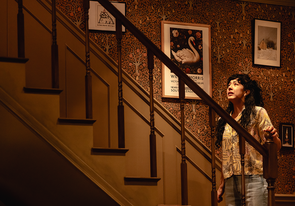 A woman stands by a wooden staircase, surrounded by floral wallpaper and framed artwork.