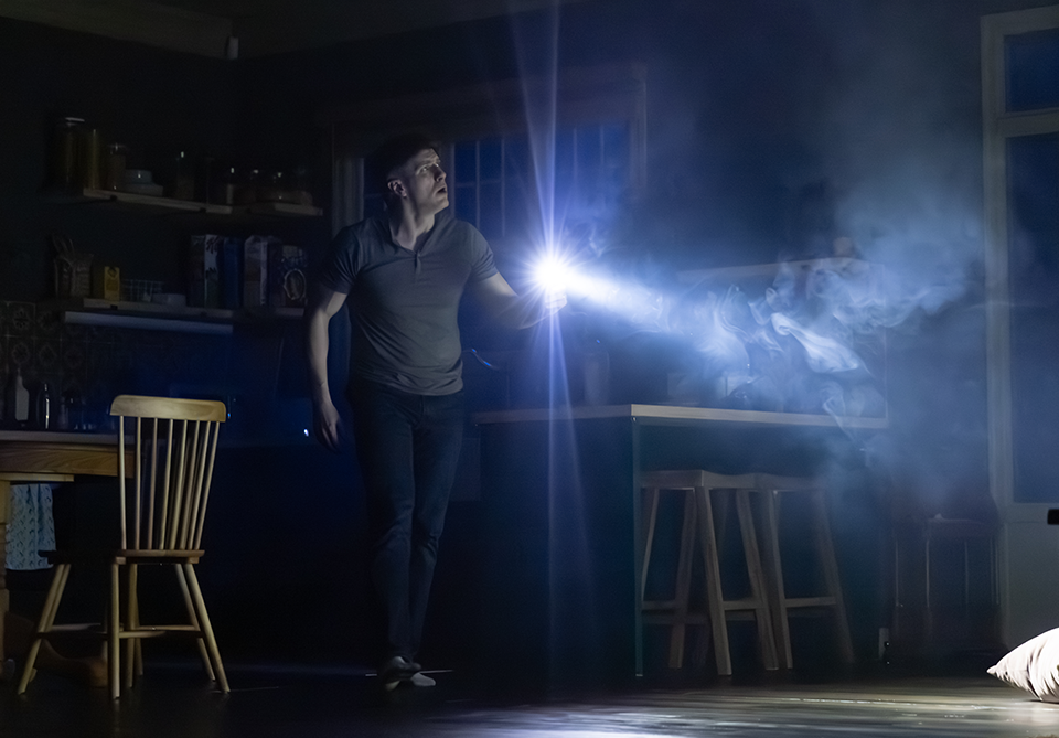 A person stands in a dark room holding a flashlight, casting a bright beam of light that reveals smoke or mist in the air.