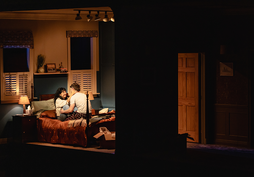 A cozy bedroom scene with two people sitting on a bed, warmly lit by a lamp with a slightly open door in the background.