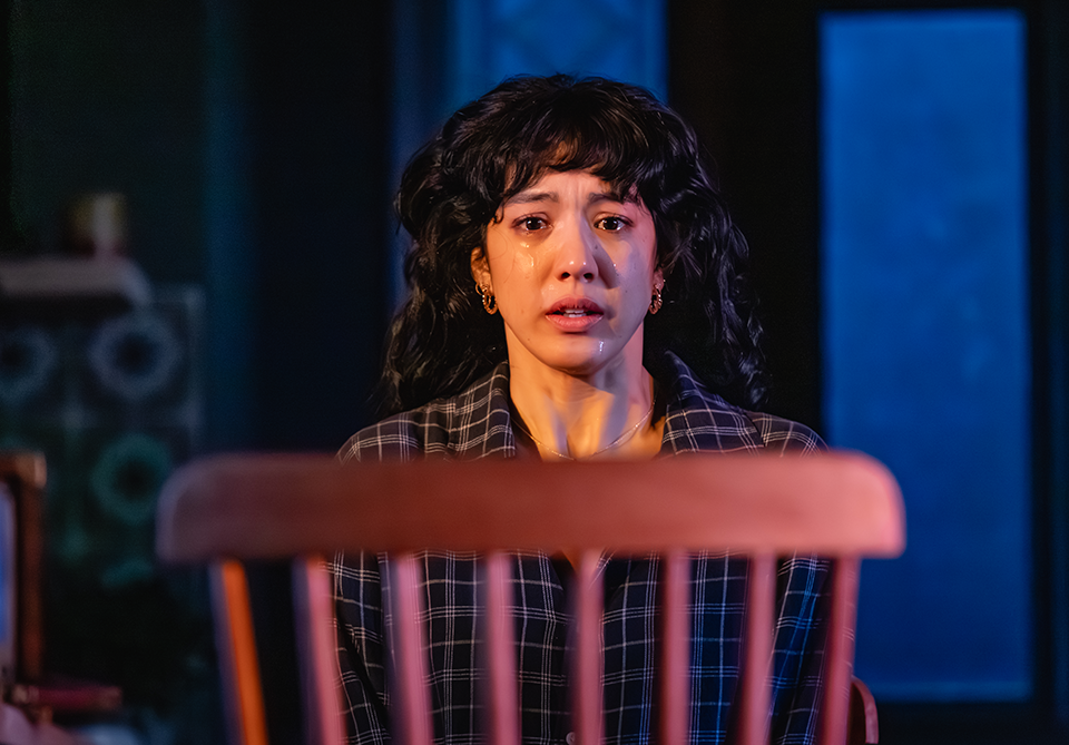 A woman with curly black hair and teary eyes looks somber behind a wooden chair.