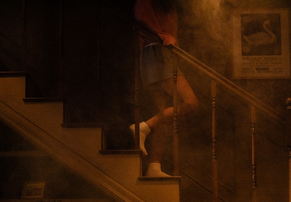 A person in white socks descending a dimly lit staircase, with a misty haze surrounding them and a poster on the wall.