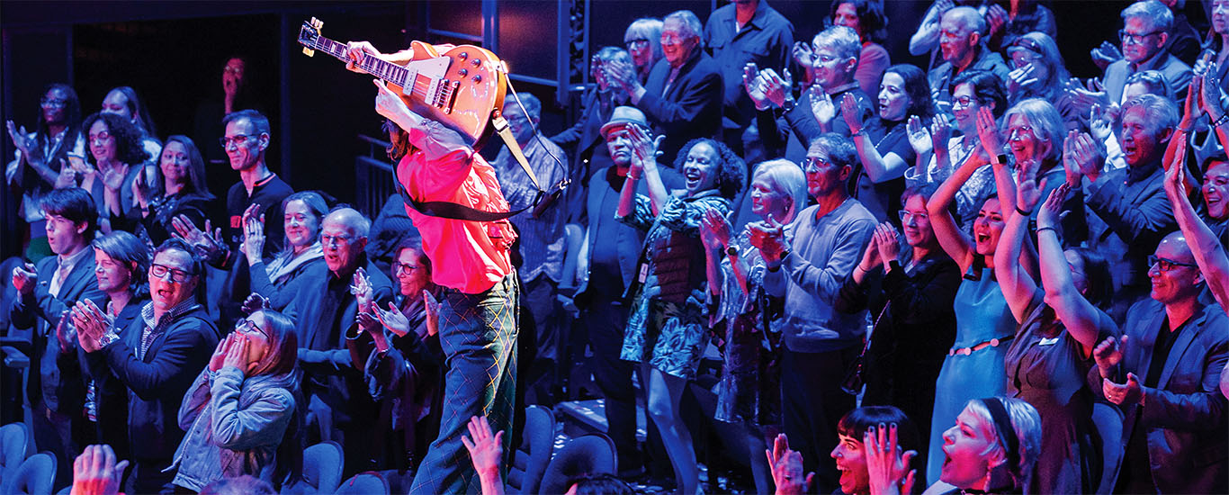 Sub Page Banner (1) A theater full of people standing and cheering looking onstage at a performer playing the guitar during the production of "Sunny Afternoon"