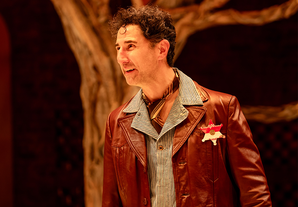 A person in a brown leather jacket and striped shirt, standing indoors with a warm-toned background and a tree trunk visible.
