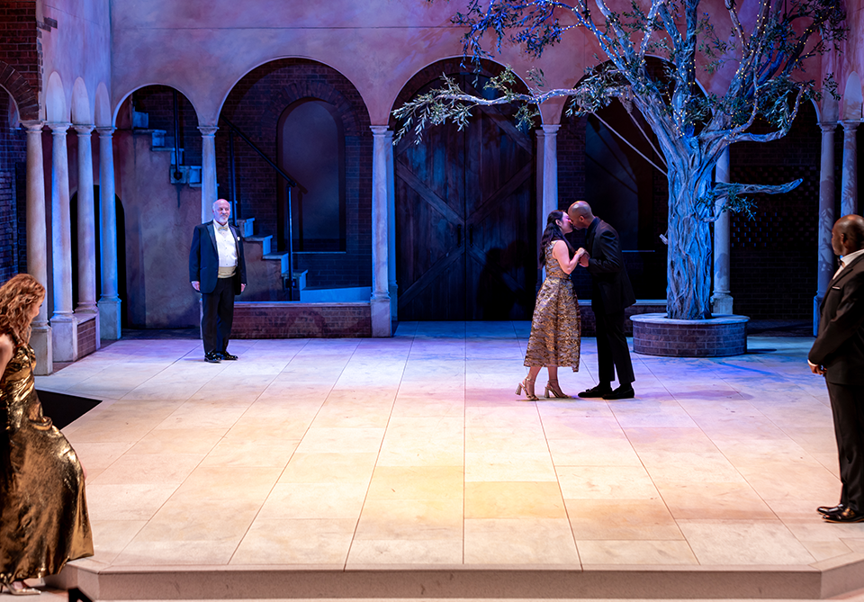 A couple kisses on stage set with columns, arches, and a lit tree.