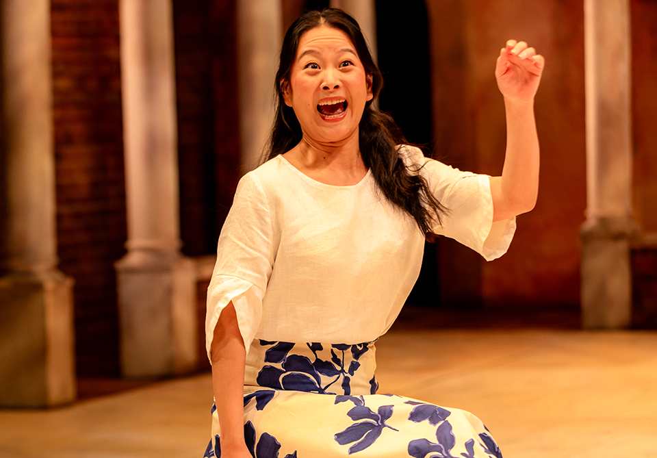 Woman in a white blouse and blue floral skirt, animatedly raising her arm in a columned setting.