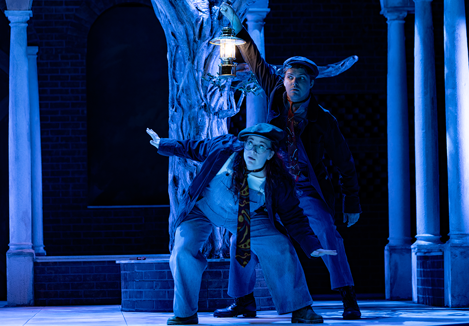 Two actors in vintage clothing, under blue light, one holding a lantern.