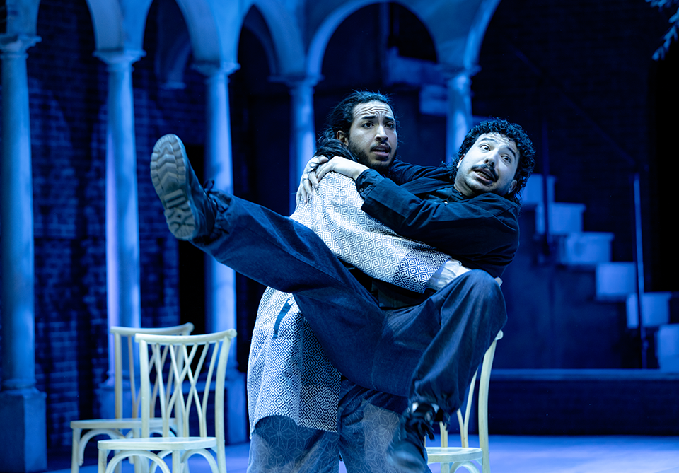 Two people on stage, one carrying the other, with blue-lit arches in the background.