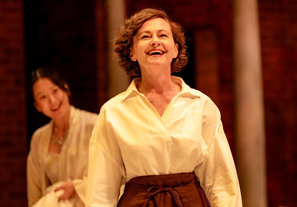 Two women smiling on a warmly lit stage, one in focus and one slightly out of focus in the background.