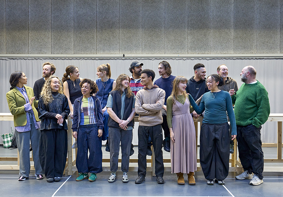 A group of fifteen people standing and conversing casually in a brightly lit rehearsal room.