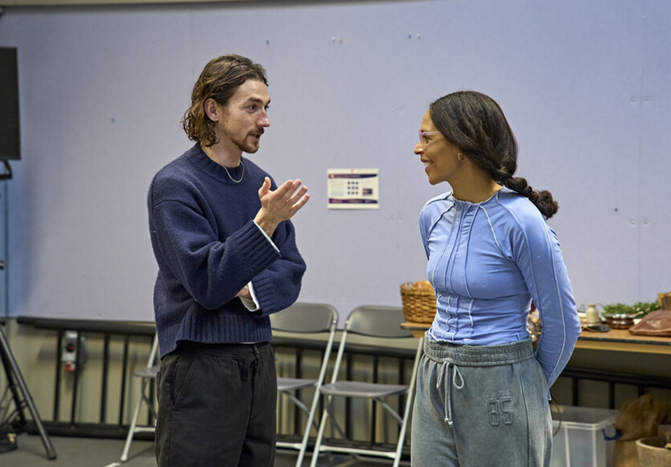 Two actors conversing in a rehearsal room with chairs and a table in the background.