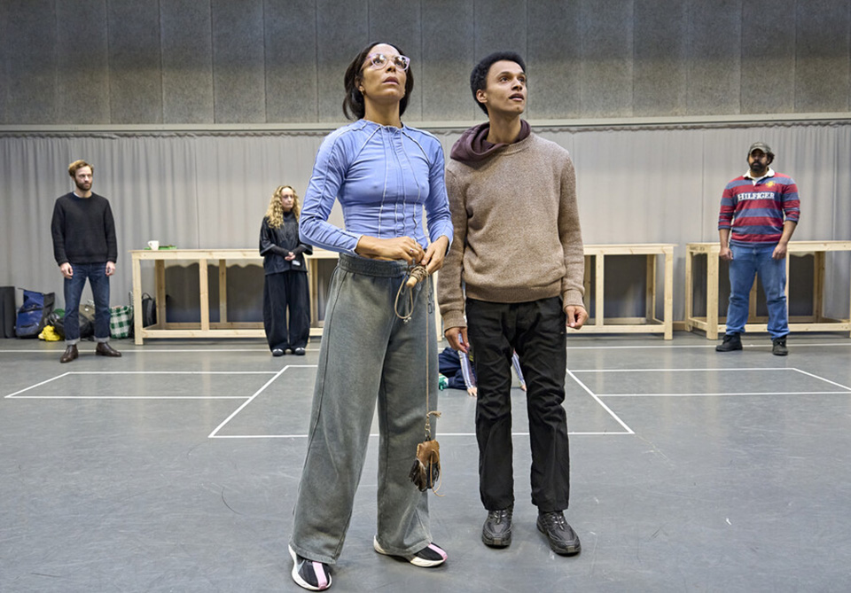 A group of people stands in a room with gray walls and floor, focusing on two individuals in the foreground looking upward.