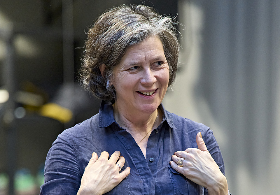 A person with short hair wearing a dark blue shirt and smiling warmly with hands lifted towards their chest.