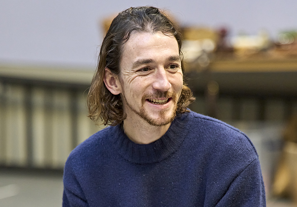 A person with shoulder-length brown hair and a beard smiling, wearing a dark blue sweater indoors.