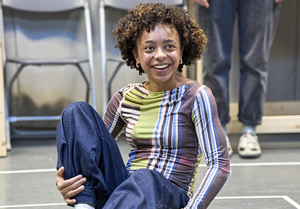 A person smiling broadly while sitting on the floor, wearing a colorful striped shirt and jeans.