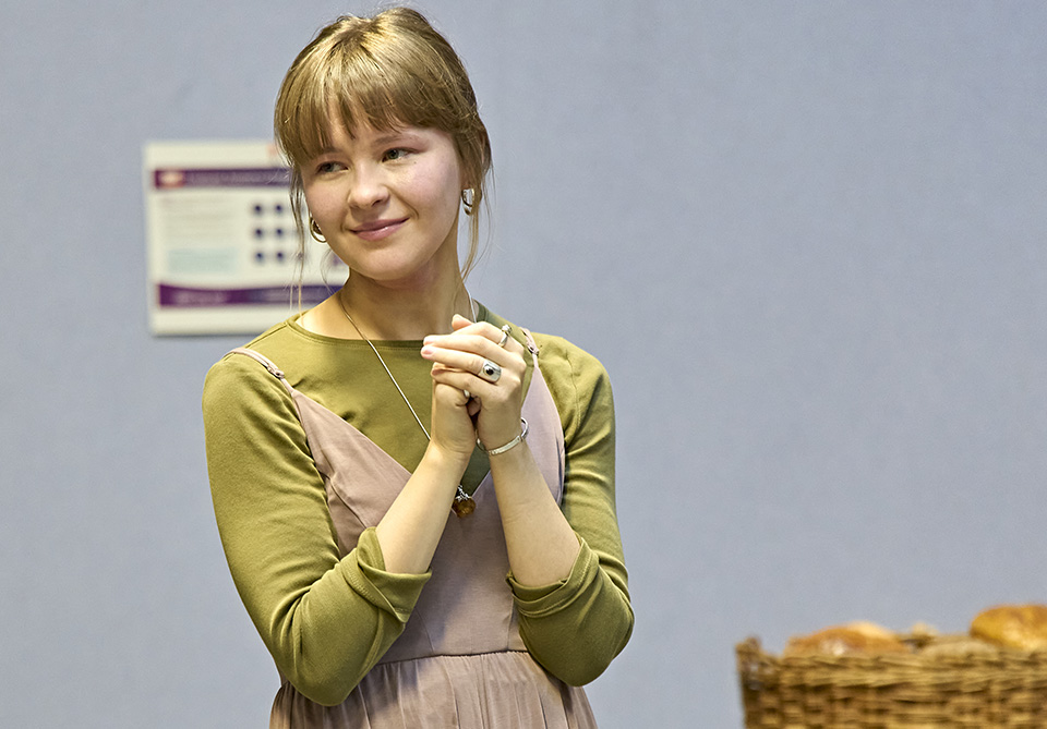 Person with blonde hair pulled back and bangs in green shirt and pink dress clasping hands, with blurred poster and basket of bread in the background.
