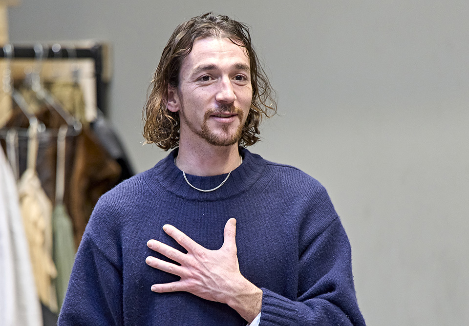 Person with long hair in a blue sweater, hand on chest, indoor background with clothes hanging on rack. 