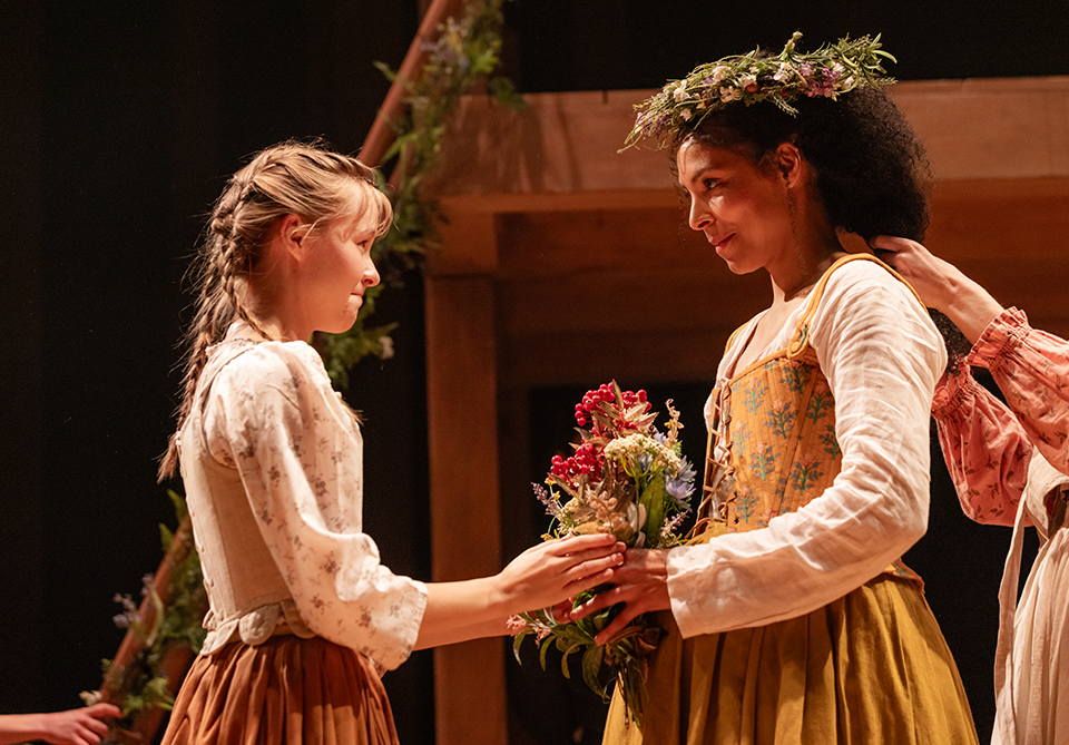 An adult woman with a wreath on her head and flowers in her hands stands across from a young woman who is receiving the flowers