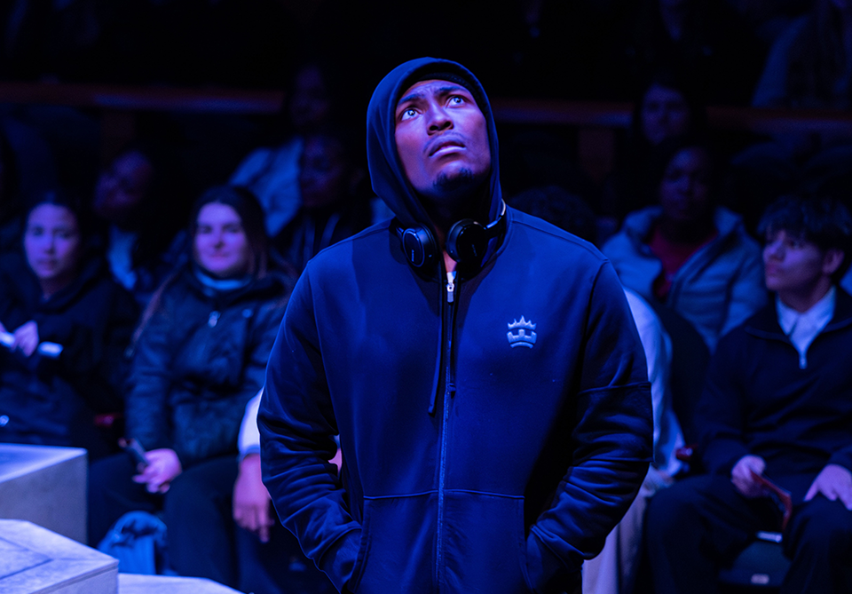 A man in a blue hoodie looks upward, with headphones around his neck, in front of a seated audience.