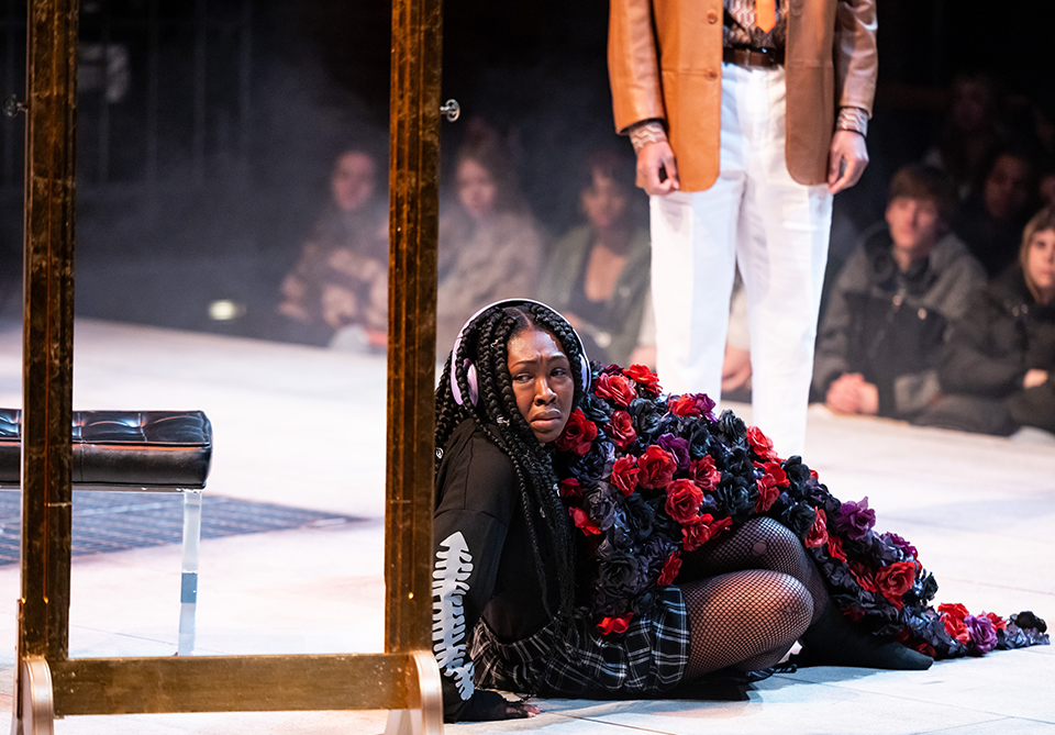 A person seated on stage, wearing headphones, with red and black flowers draped over them, next to a standing figure.