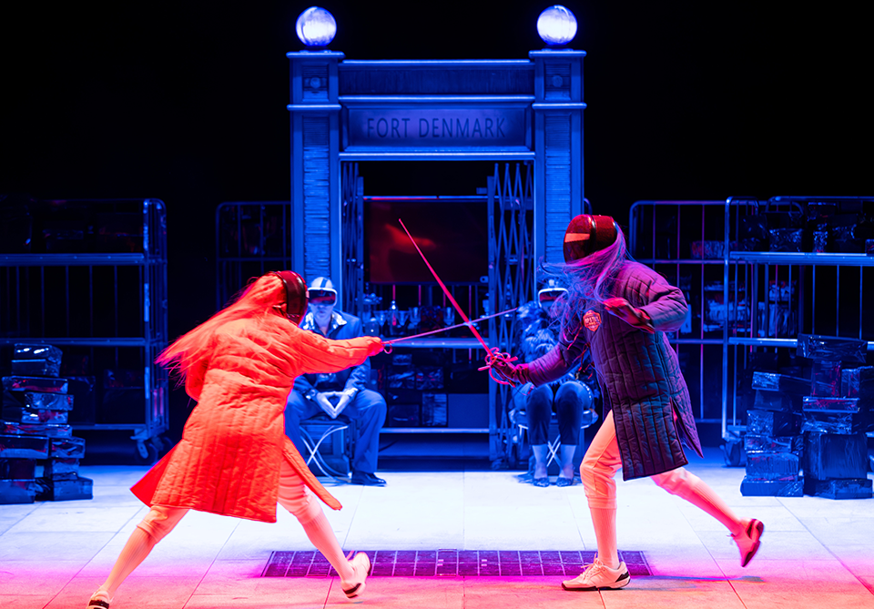 Two fencers duel on stage under red and blue lighting in front of a gate marked "FORT DENMARK."