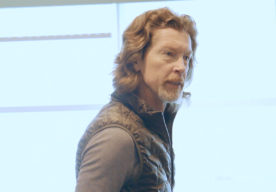 Person with medium-length wavy hair and a beard in a quilted vest stands in front of bright windows.