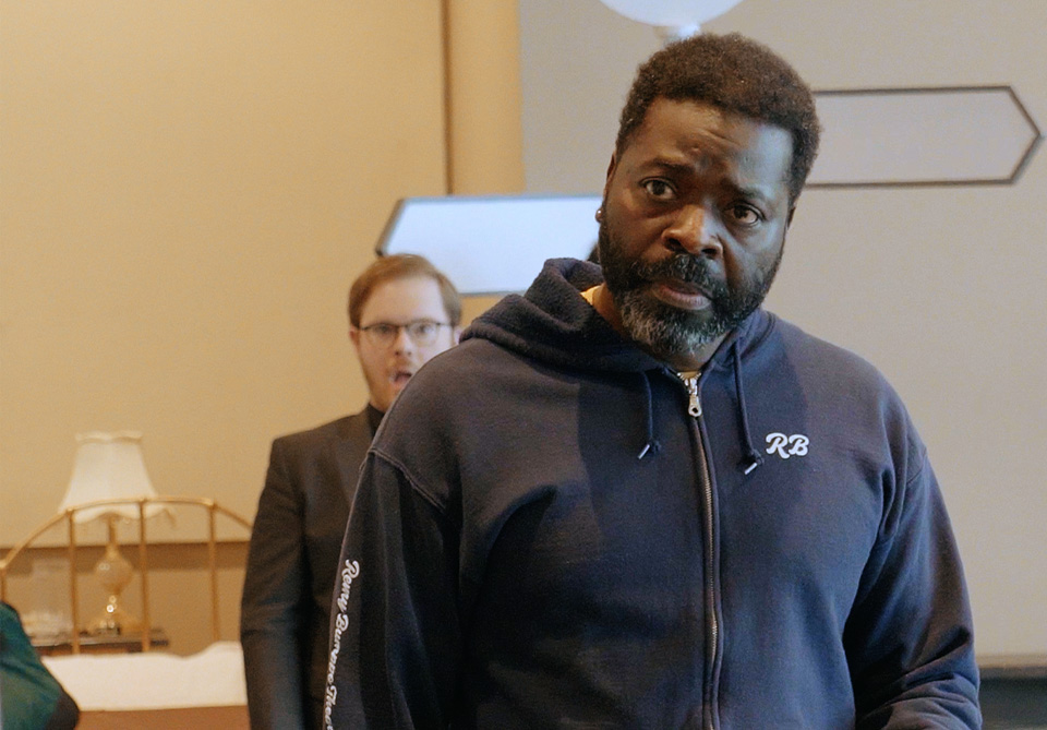 A man in a navy blue hoodie stands in the foreground with a serious expression. Another man in glasses is visible in the background.