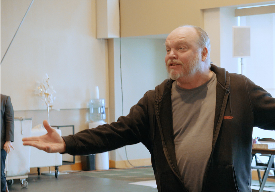 A man in a dark hoodie and gray T-shirt stands indoors with arms outstretched, gesturing expressively.
