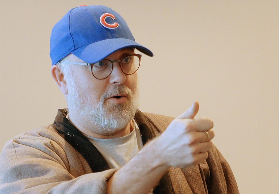 A person with a beard wearing a blue baseball cap and glasses, giving a thumbs-up gesture.