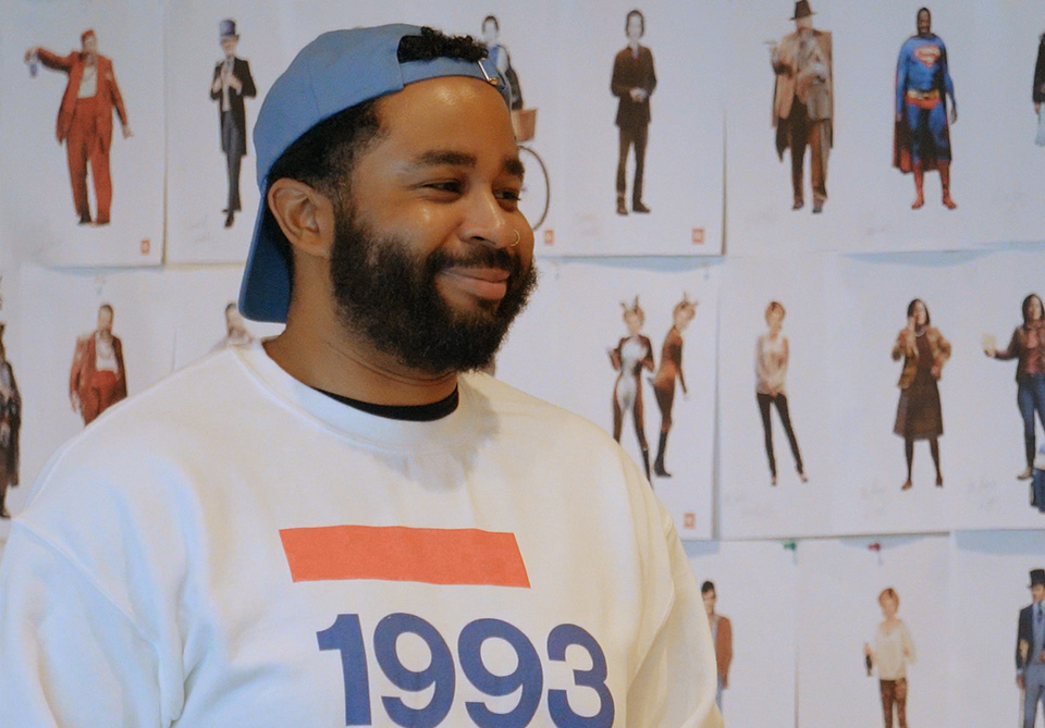 Person wearing a "1993" sweatshirt with character and costume design illustrations on a wall in the background.