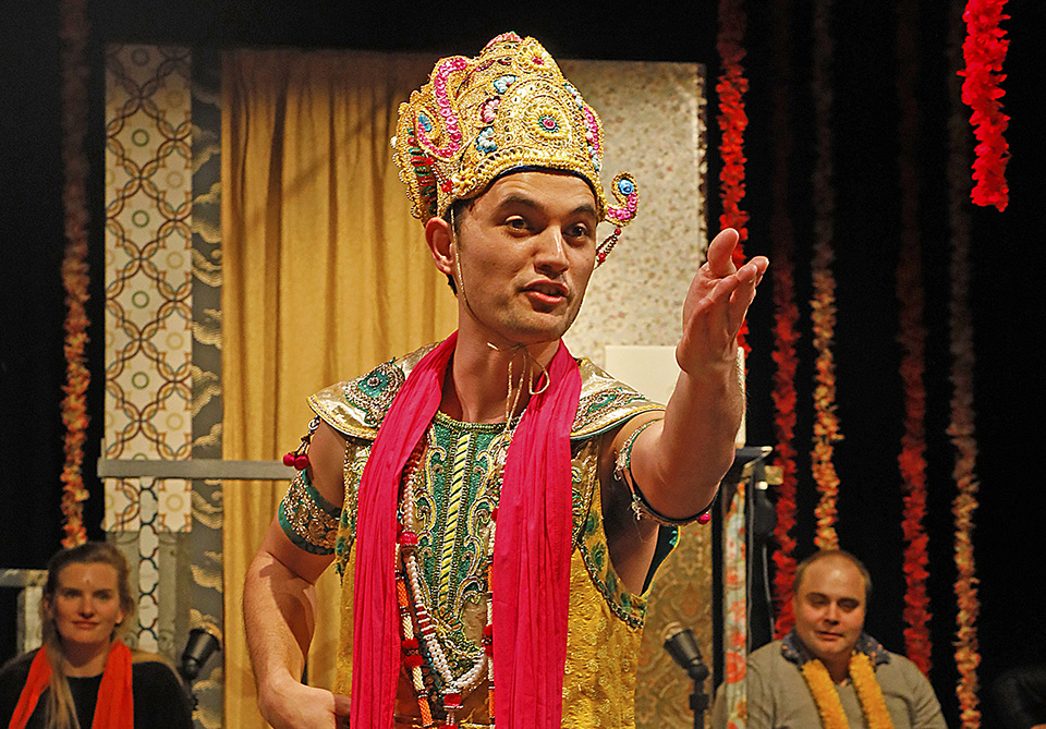Man in colorful costume performing on stage with ornate headdress and extended hand.