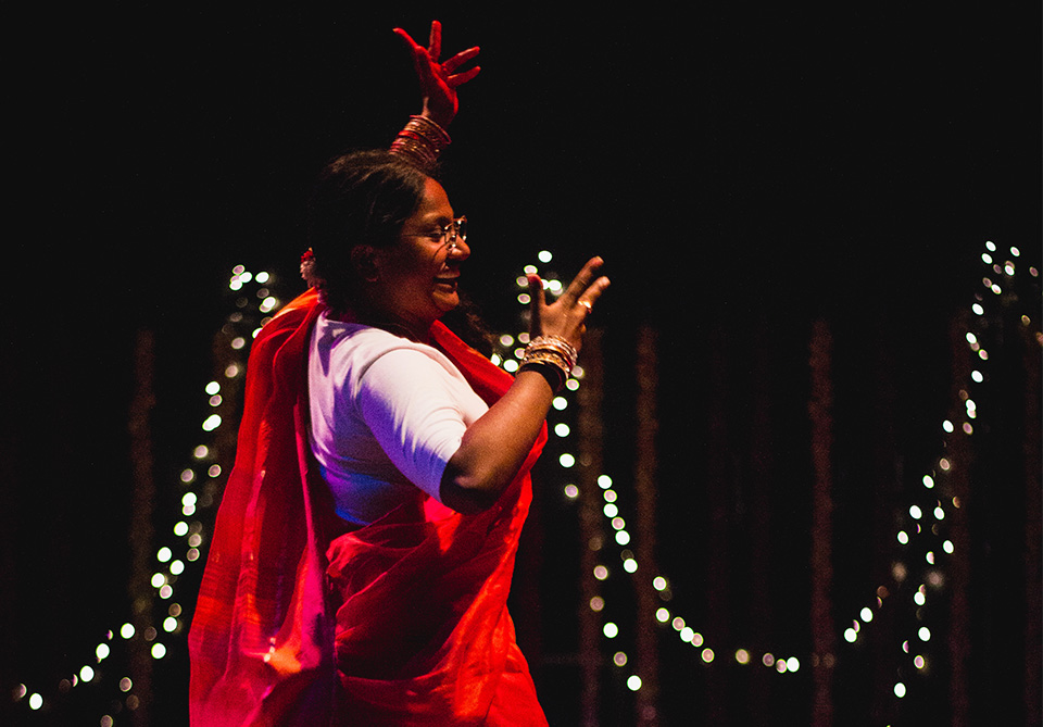 A person dances in a red sari with a dark background and string lights.