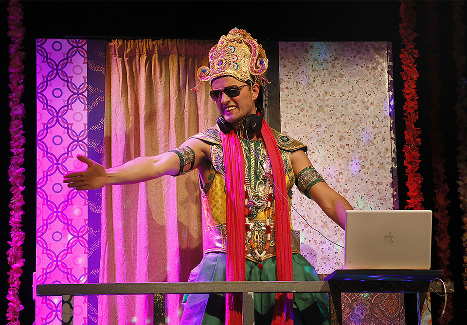 Person in a traditional costume and sunglasses behind a DJ booth with a laptop in a colorful setting.