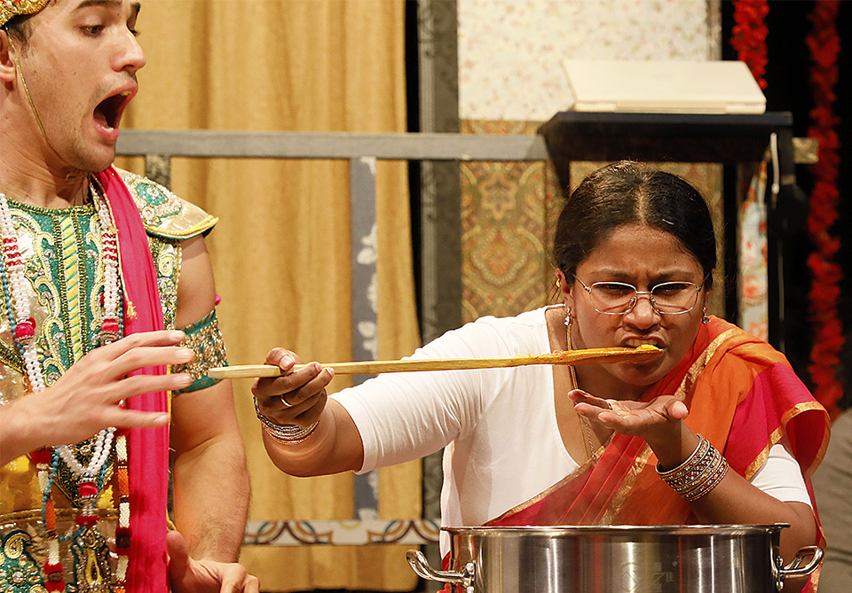 Two people in traditional attire on stage; one is tasting food from a long spoon.