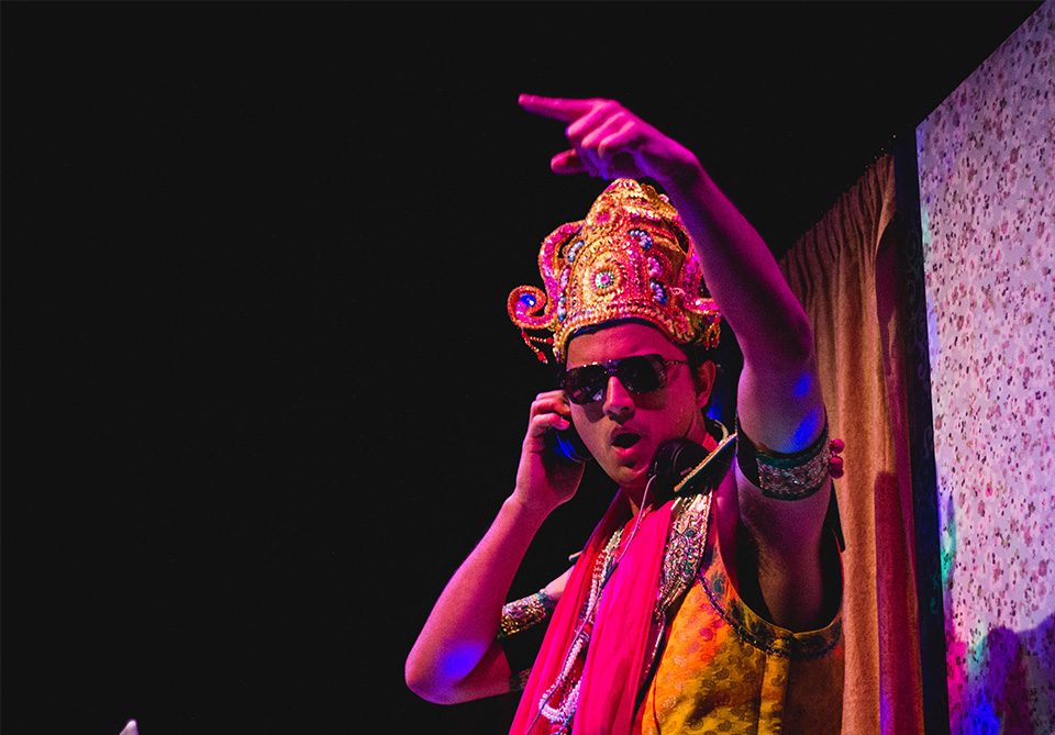 Person in colorful traditional attire with a jeweled headpiece and sunglasses on stage.