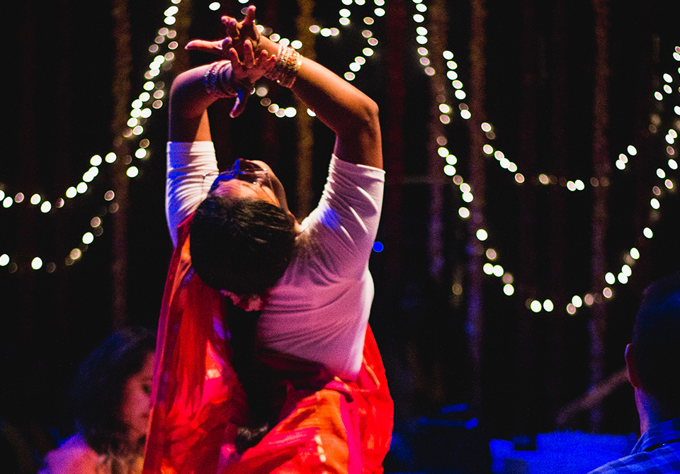 A dancer in a red and orange saree performs under twinkling lights.
