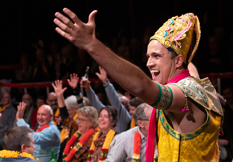 Man in a vibrant costume with a headdress gesturing energetically in front of an engaged audience wearing colorful garlands.