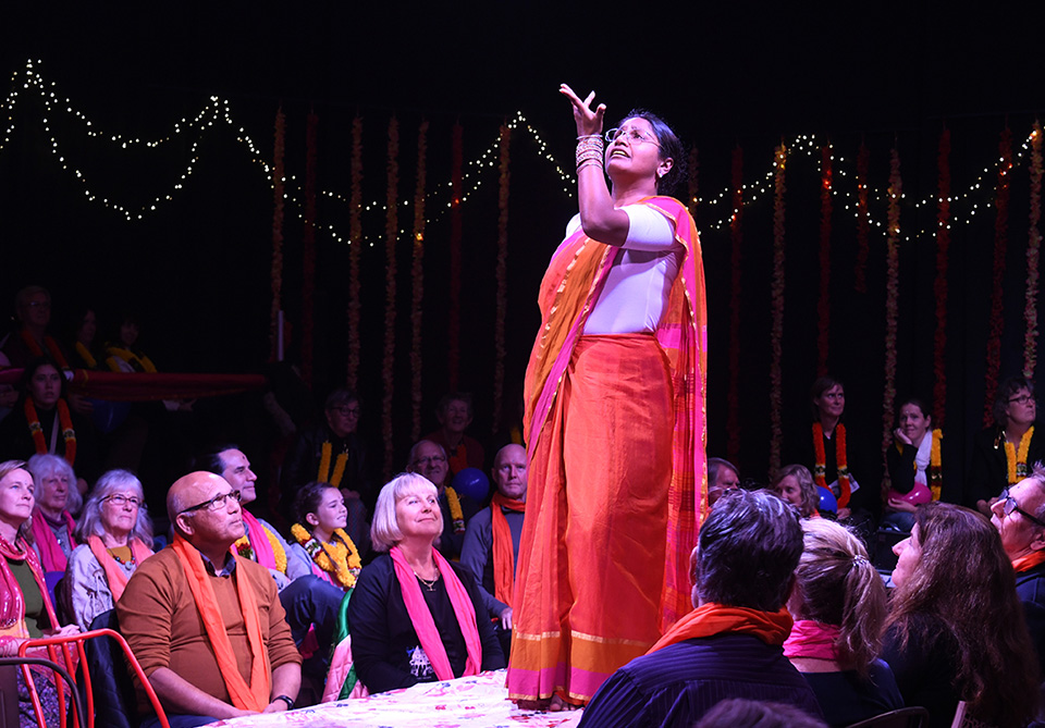 A performer in an orange and pink saree gestures on stage surrounded by a seated audience with twinkling lights in the background.