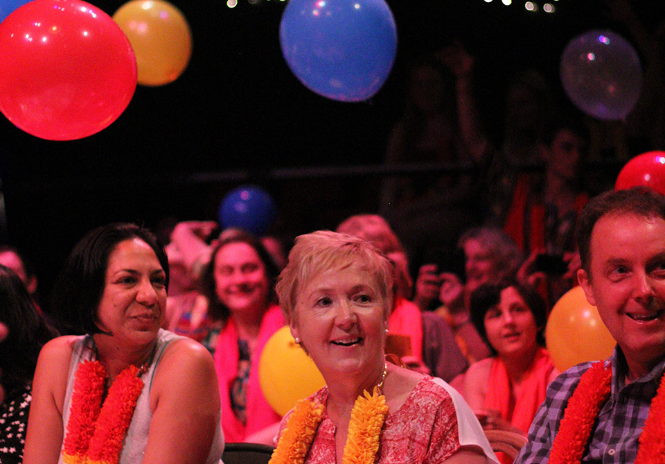A happy group of people at an indoor event with colorful leis and floating balloons.
