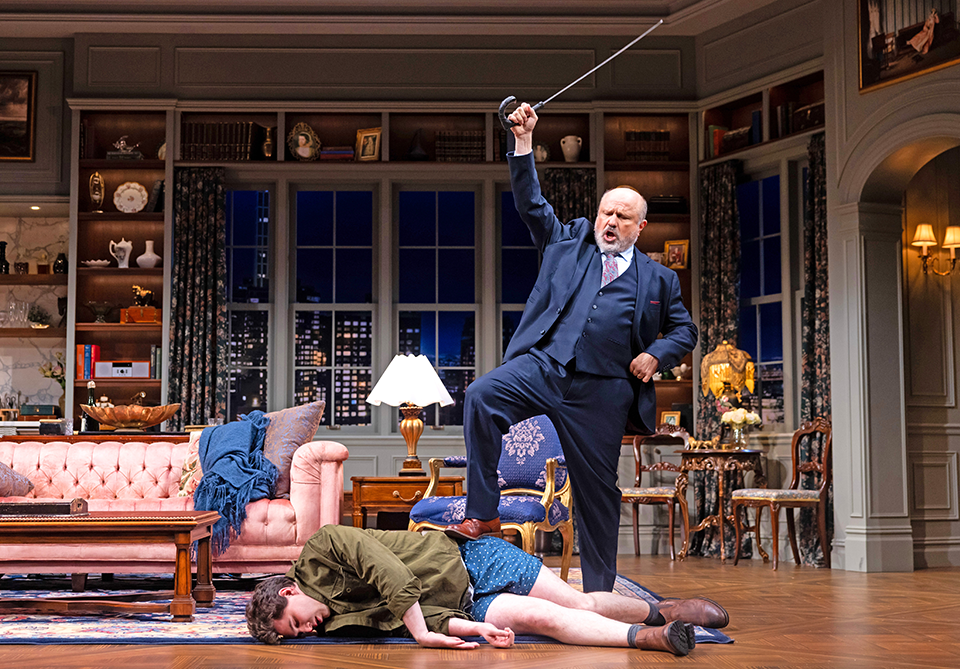 A dramatic scene with a man in a suit holding a sword over another man lying on the floor in a richly furnished living room.