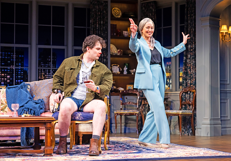 A man in casual attire sits on a chair while a woman in a blue suit joyfully gestures in a cozy, elegant living room.