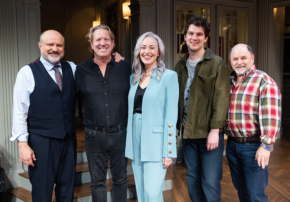 A group of five people stand closely together, smiling warmly. They're indoors on a wooden floor, dressed in a mix of formal and casual attire, creating a welcoming and friendly atmosphere.