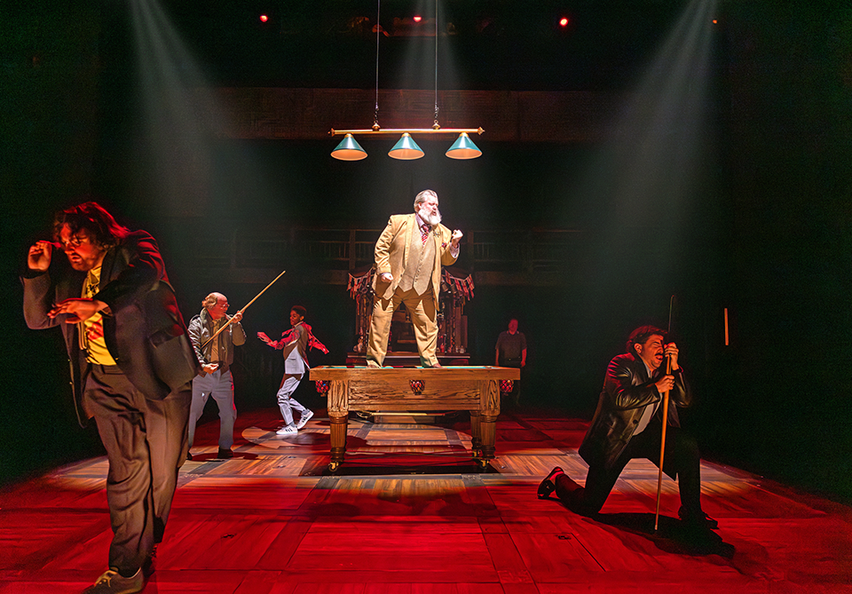 A stage scene with five performers, one standing on a pool table, surrounded by others in dramatic poses under overhead lights.