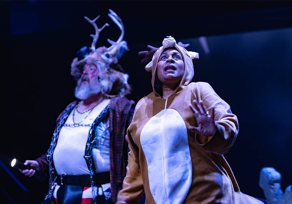 Two people in a theatrical scene; one with a white shirt and suspenders with antlers on his head, the other in a reindeer onesie.