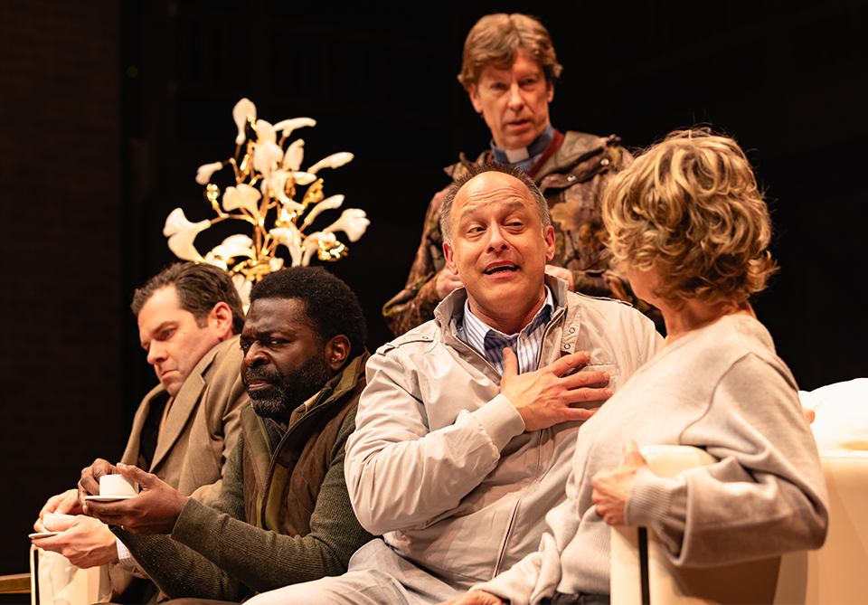 A group of five people in a theatrical setting with warm lighting and a decorative floral backdrop.