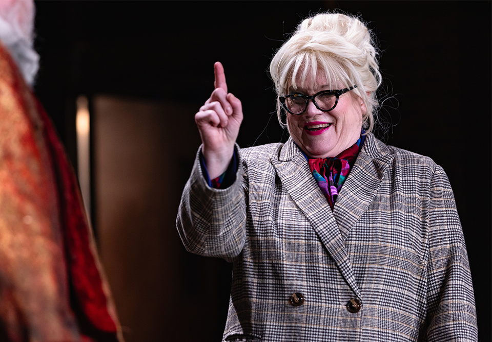A person with blonde hair in an updo, wearing glasses and a checkered blazer, smiles with a raised finger.