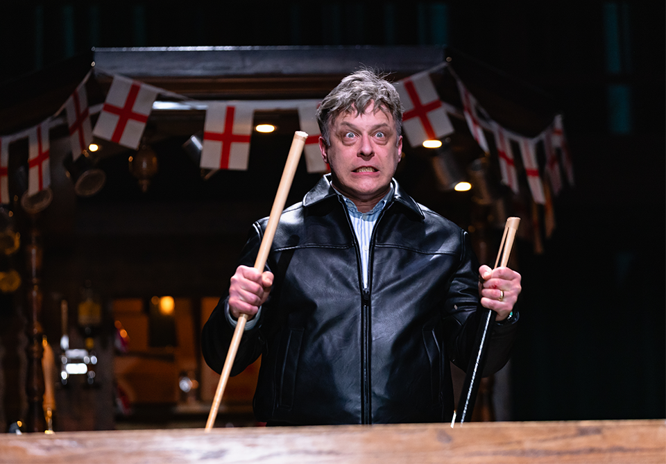 Person in a black leather jacket holds two sticks, with flags hanging in the background.